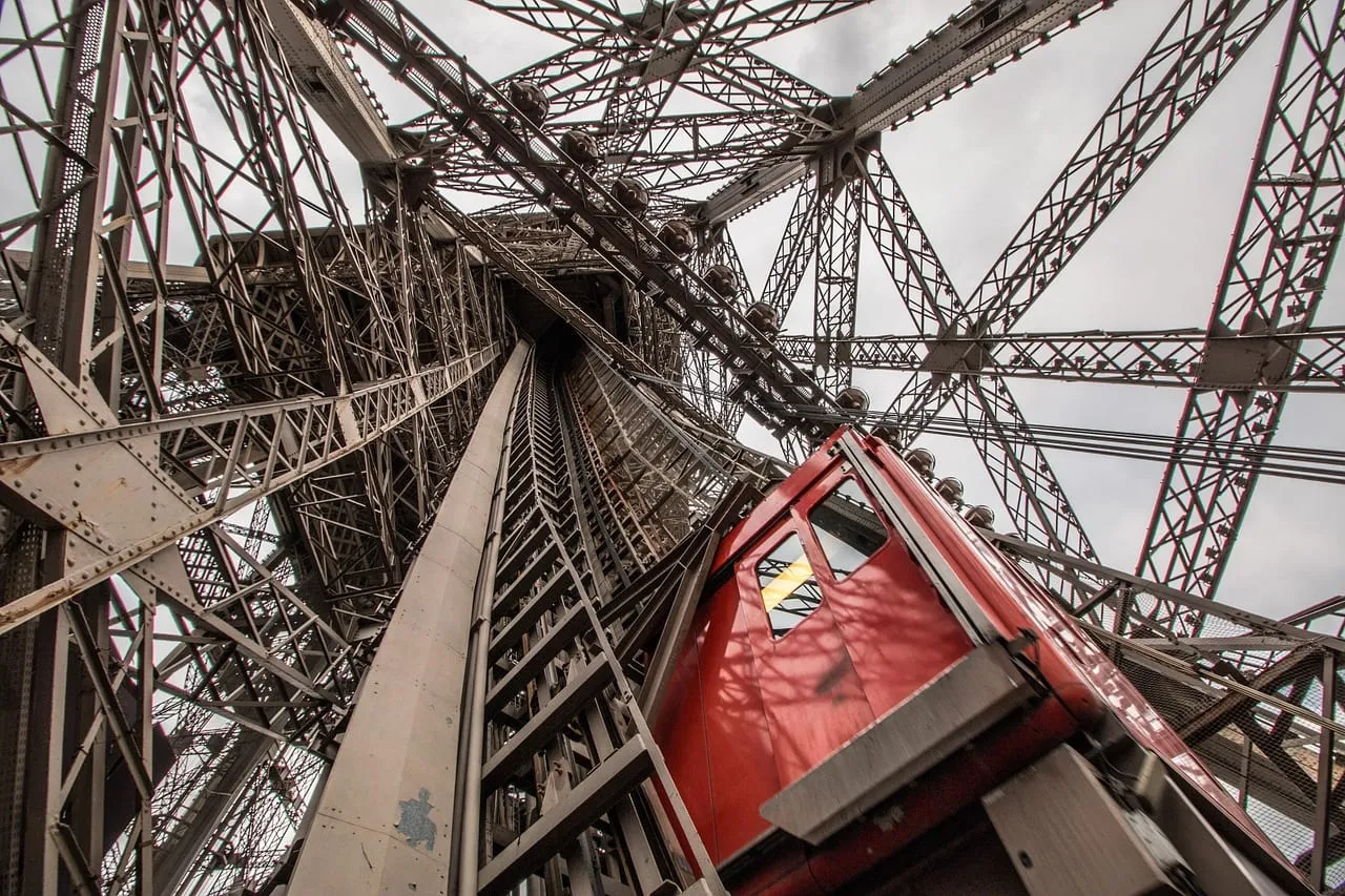 Une cabine d’ascenseur rouge gravissant la charpente métallique de la tour
