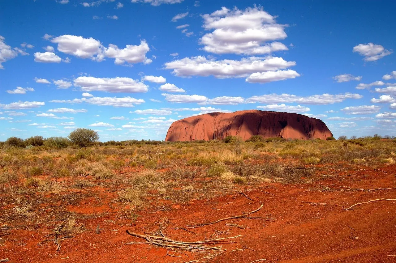 L’Uluru à l’horizon, au-delà d’une terre rouge parsemée de buissons
